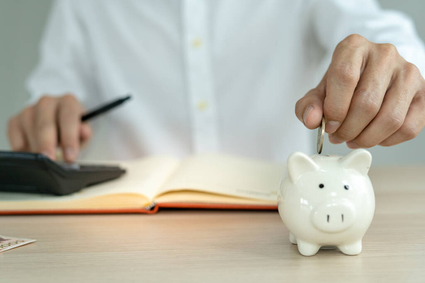 The man is putting two coins into his savings each month. A pig piggy bank holder and is show to the front as a symbol of savings. monthly savings concept. - Photo, Image