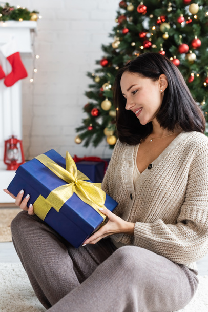 happy brunette woman looking at blue gift box with yellow ribbon near blurred christmas tree - Photo, Image