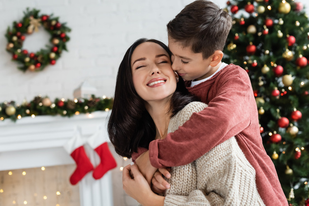 мальчик обнимает счастливую мать возле размытой рождественской елки дома - Фото, изображение