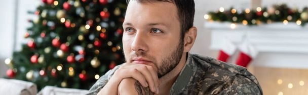 pensive military man holding hands near face and looking away near christmas tree, banner - Фото, зображення