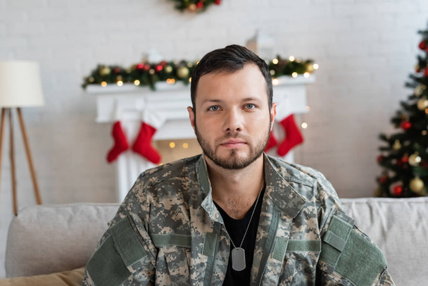 sérieux homme militaire non rasé regardant la caméra près de la cheminée avec décor de Noël sur fond flou - Photo, image