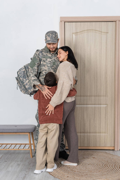 άντρας με καμουφλάζ αγκαλιάζει γιο και ευτυχισμένη σύζυγο κοντά στην πόρτα εισόδου στο σπίτι - Φωτογραφία, εικόνα