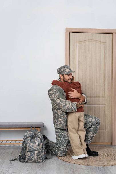 gelukkig militair man omarmen zoon in de buurt van rugzak en ingang deur thuis - Foto, afbeelding
