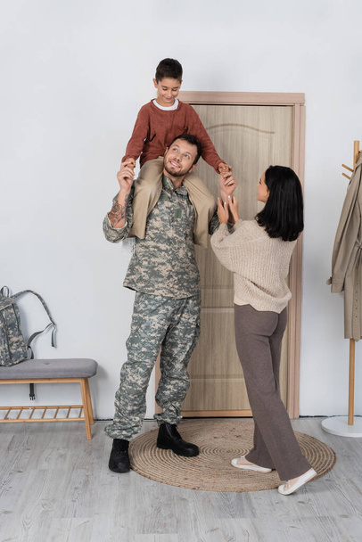 smiling boy piggybacking dad in military uniform near wife and entrance door at home - Foto, Imagem
