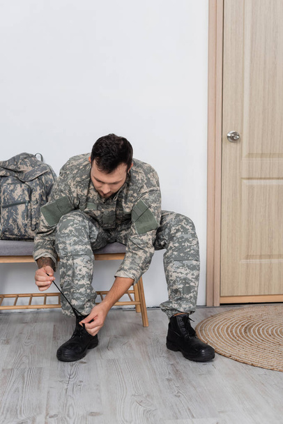 man in camouflage sitting near backpack and unlacing military boot at home - Photo, Image