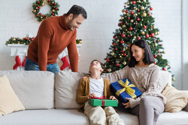 happy boy looking at dad while sitting on couch with christmas presents and smiling mom - 写真・画像