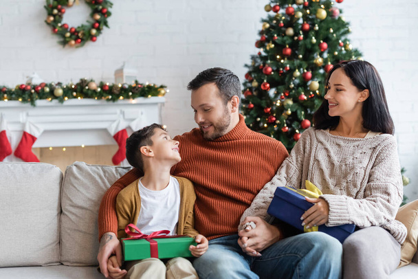 happy man holding hands with wife and looking at cheerful son on sofa near gift boxes and blurred christmas tree - Foto, imagen
