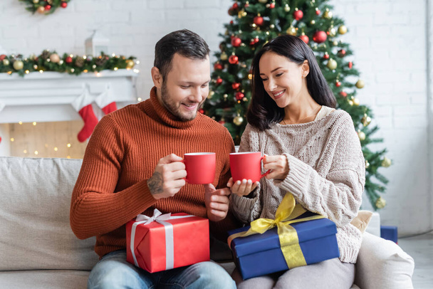 cheerful couple with gift boxes and cups of warm cocoa sitting near christmas tree on blurred background - 写真・画像