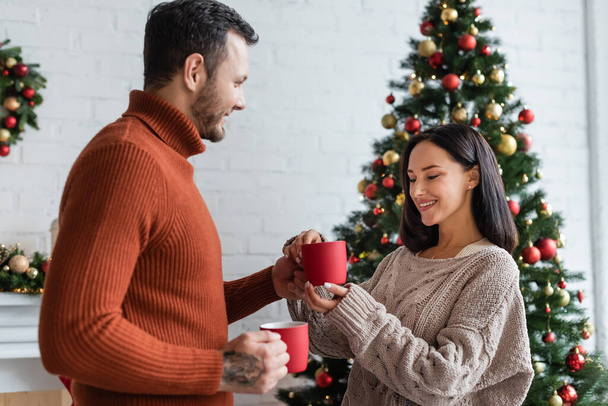smiling man giving cup of warm cocoa to pleased wife near decorated christmas tree - Foto, Imagen