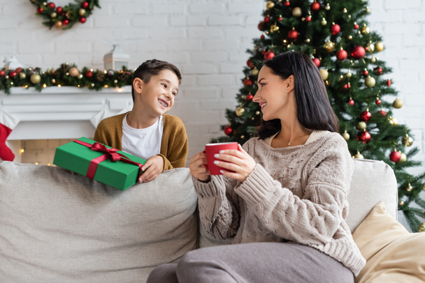 cheerful boy with christmas present near mom sitting on couch with cup of warm cocoa - Foto, Imagen