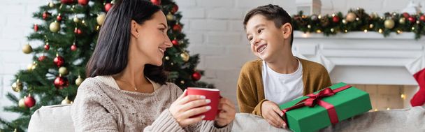 femme heureuse avec tasse de cacao chaud regardant fils gai avec cadeau de Noël, bannière - Photo, image