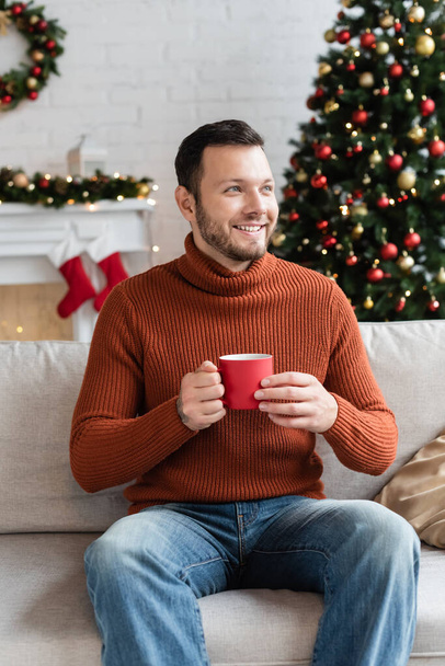 happy man with cup of cocoa looking away on sofa near christmas tree on blurred background - Photo, Image