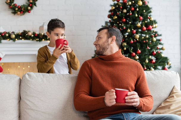 cheerful man sitting on couch and looking at son drinking warm cocoa near decorated christmas tree - Фото, изображение