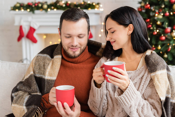 smiling couple with cups of cocoa sitting under warm blanket in living room with blurred christmas decor - Photo, Image