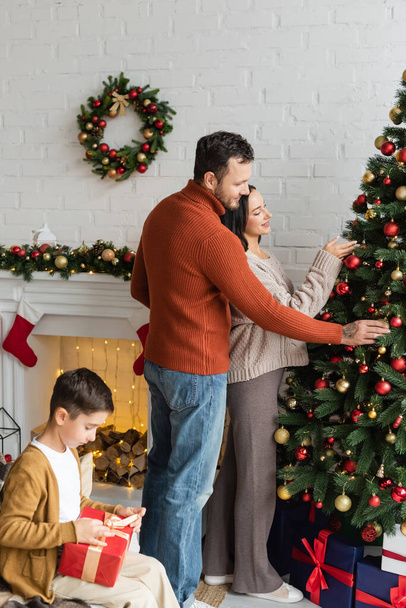 Çocuk Noel hediyesini paketlerken, ebeveynler evde yeşil ladin süsleme yapıyor. - Fotoğraf, Görsel