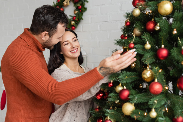cheerful couple decorating green spruce with christmas balls in living room - Foto, immagini