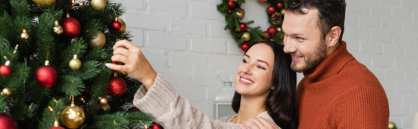 szczęśliwy człowiek uśmiechnięty w pobliżu brunetka żona dekorowanie zielony sosna z bożonarodzeniowe kulki, baner - Zdjęcie, obraz