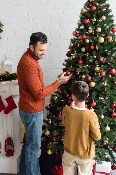 usmívající se muž při pohledu na syna při zdobení vánoční stromeček s ozdoby doma - Fotografie, Obrázek