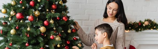 брюнетка улыбается рядом с сыном украшая зеленую ель рождественскими шарами, баннер - Фото, изображение