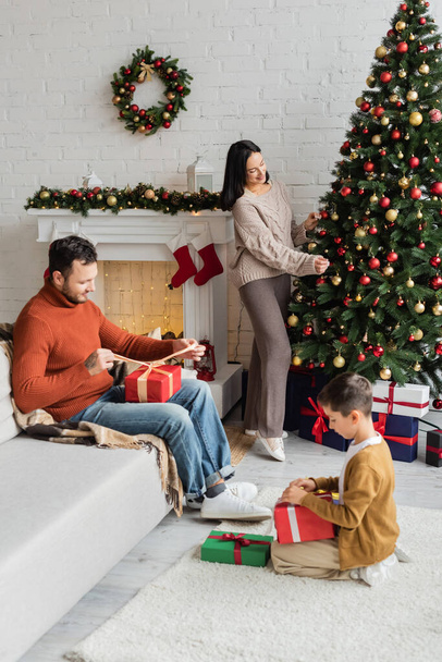 happy woman decorating christmas tree near father and son packing presents in living room with fireplace - Φωτογραφία, εικόνα
