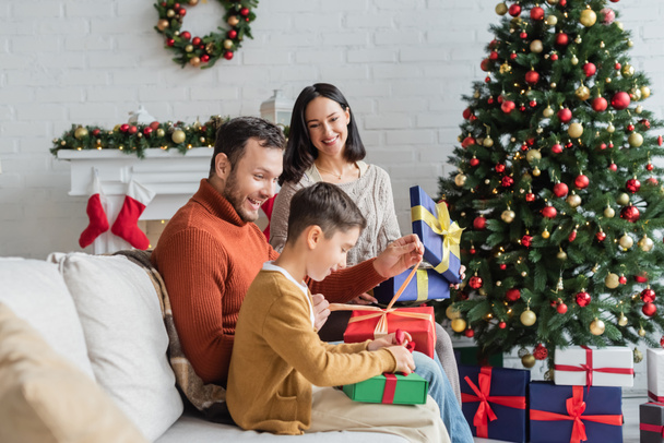 joyful family packing gift boxes on couch near christmas tree and decorated fireplace - Foto, imagen