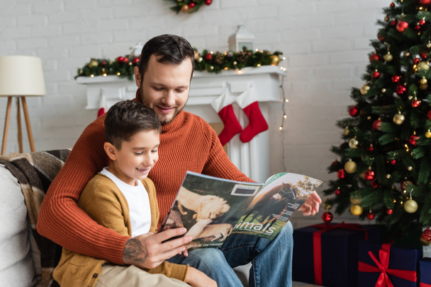 smiling dad and son reading magazine in living room with christmas decor - Photo, Image