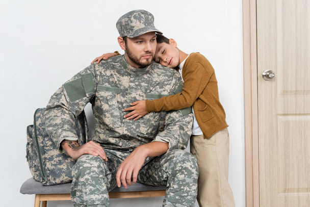 kid with closed eyes embracing serious dad in camouflage sitting near backpack - Photo, Image