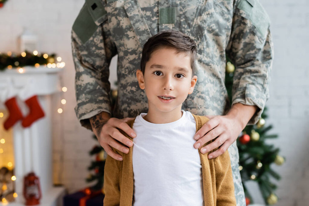 軍の制服を着た男が家でカメラを見ている息子の肩を抱きかかえて - 写真・画像