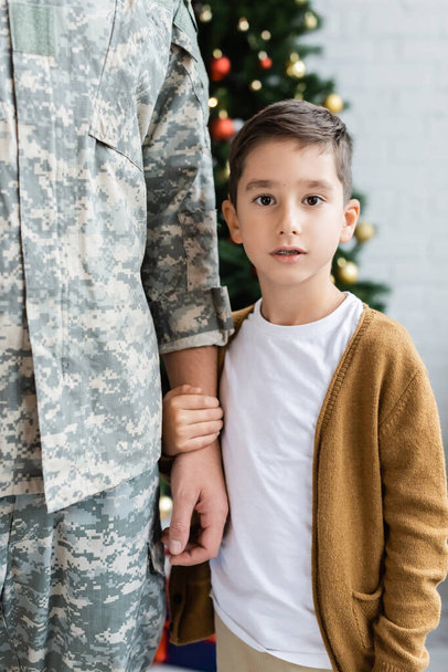 kid looking at camera while holding hand of dad in camouflage at home - Foto, Bild