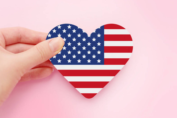 Mão feminina segura bandeira de papel americana em forma de coração isolado sobre fundo rosa, dia 4 de julho independência da América, celebração, cartão postal, cartão de saudação - Foto, Imagem