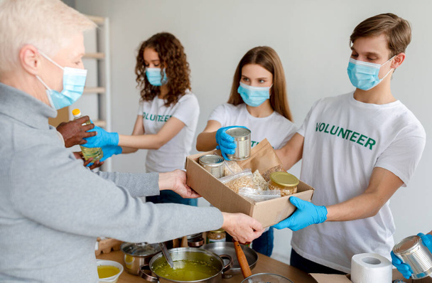 Grupa młodych wolontariuszy w maskach dających żywność w pudełkach kartonowych jako darowiznę dla osób ubogich i starszych. Millenialsi pracujący w ośrodku charytatywnym - Zdjęcie, obraz