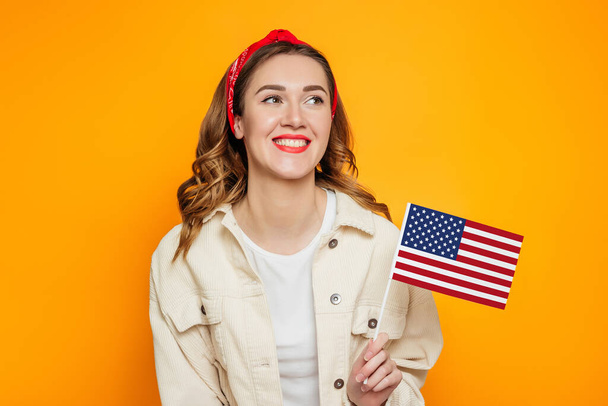 Happy young woman with red lipstick holds a small american flag and smiles isolated over orange background, girl holding USA flag, 4th of july independence day, copy space - Photo, image