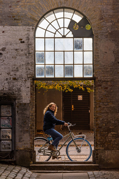 Copenhague, Dinamarca Una ciclista maniobra una bicicleta sobre un paseíto en la isla de Reffen bajo un pasillo arqueado. - Foto, imagen