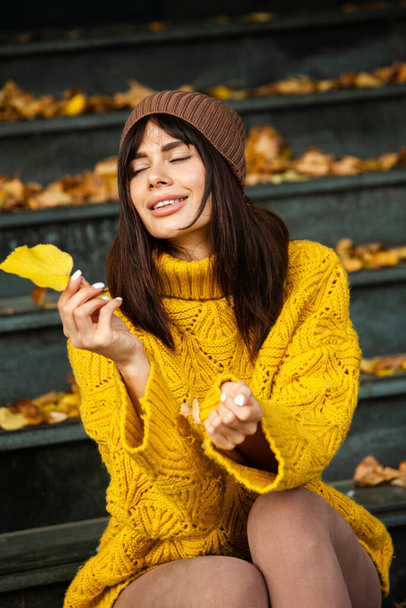 Beautiful European brunette dressed in a yellow woolen sweater and hat outside. The beautiful girl who wears thick stylish autumn clothes in cool weather. - Fotografie, Obrázek