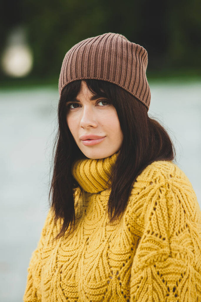 Beautiful European brunette dressed in a yellow woolen sweater and hat outside. The beautiful girl who wears thick stylish autumn clothes in cool weather. - Photo, Image