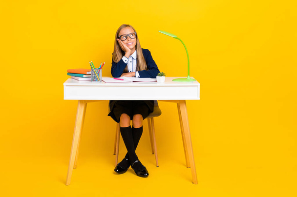 Full photo of dreamy schoolgirl with blond hair wear blue blazer sitting at desk look empty space isolated on yellow color background. - 写真・画像