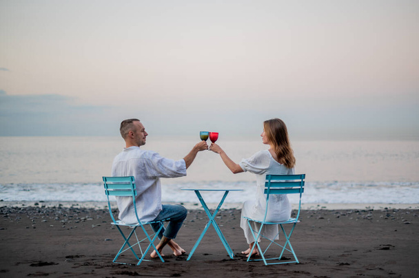 Young couple sitting on a beach on sunset and drinking wine out of glass. High quality photo. Dressed in white tops and jeans. - Valokuva, kuva