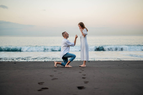 Yong uomo che fa una proposta di matrimonio su una spiaggia al tramonto ad una giovane bella donna. In piedi in ginocchio. Macchie sulla sabbia. L'uomo sta tenendo un anello di fidanzamento . - Foto, immagini