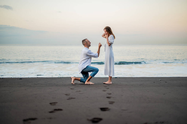 Yong man doet een huwelijksaanzoek op een strand bij zonsondergang aan een jonge mooie vrouw. Op de knie staan. Vlekken op het zand. De man heeft een verlovingsring vast. . - Foto, afbeelding