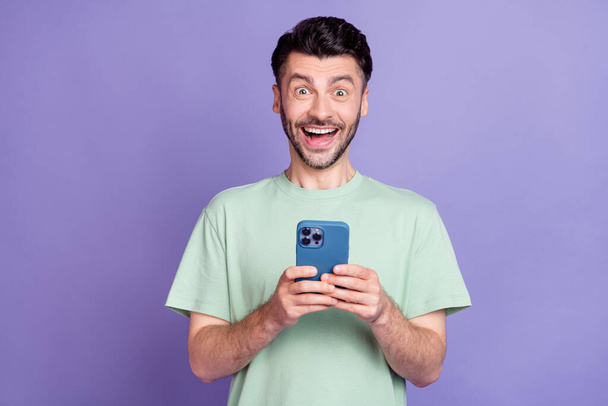Photo portrait of nice young guy hold telephone excited unbelievable news wear trendy gray garment isolated on violet color background. - Zdjęcie, obraz