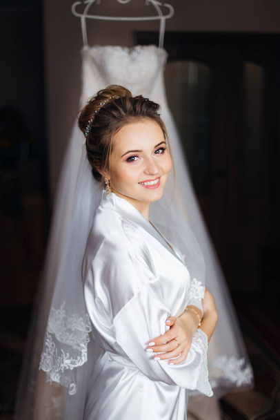 bride in white bathrobe with make up and bridal veil crossed arms and look at camera. wedding dress in the background.  - 写真・画像