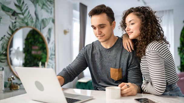Two people young adult couple man and woman husband and wife or boyfriend and girlfriend in the kitchen with laptop computer checking online content shopping online or have video call morning routine - Φωτογραφία, εικόνα