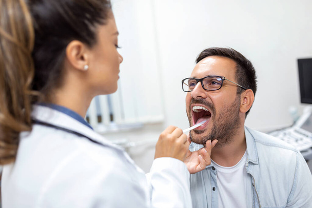 A young man sits on an exam table across from his doctor. The doctor reaches forward with a tongue depressor as the man looks up and sticks out his tongue. - Photo, Image