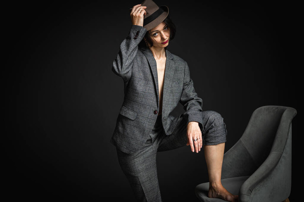 Retrato de cerca de una mujer elegante, bonita y sexy con un traje gris, escondido por el sombrero de un hombre negro. Belleza modelo de moda con labios rojos y maquillaje de ojos sobre un fondo gris oscuro - Foto, imagen