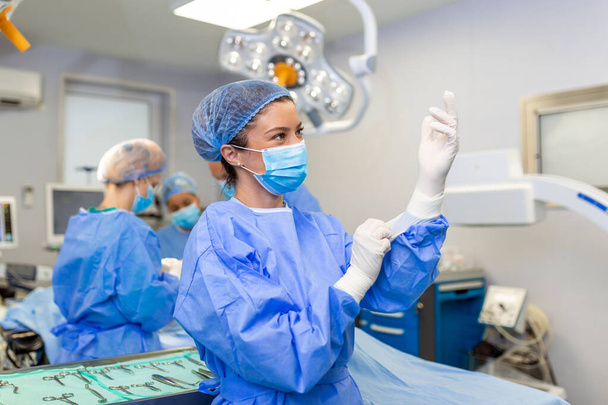 Retrato de bela médica cirurgiã colocando luvas médicas em pé na sala de operação. Cirurgião na moderna sala de cirurgia - Foto, Imagem