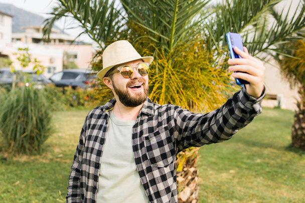 Мужчина фотографирует селфи на фоне пальм - Счастливый парень тысячелетия наслаждается летним отдыхом в городе - Молодежь и технологии. - Фото, изображение