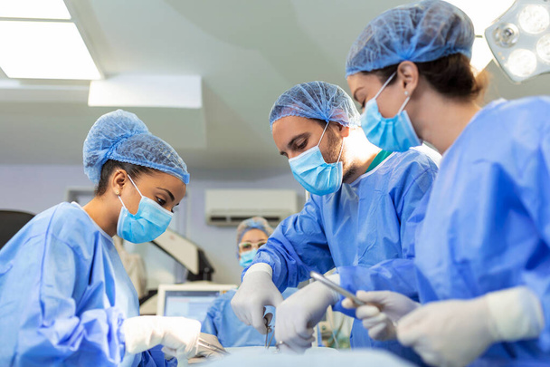 Многонациональные медицинские работники, выполняющие операции на пациенте в операционном зале. Медицинские коллеги, работающие в отделении скорой помощи больницы. Они в халатах. - Фото, изображение