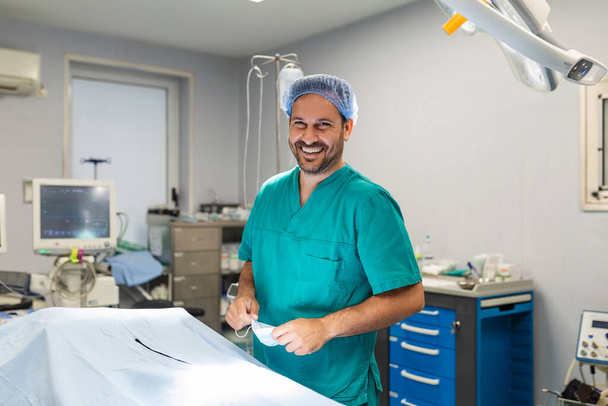 Porträt eines männlichen Chirurgen im Operationssaal, der in die Kamera blickt und OK-Geste zeigt. Arzt in Peelings und medizinischer Maske im modernen Operationssaal eines Krankenhauses. - Foto, Bild