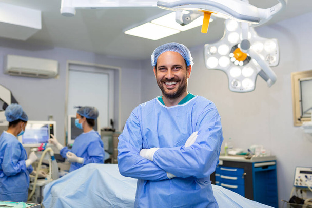 Портрет мужчины-хирурга, стоящего со скрещенными руками в операционной в больнице. Командные хирурги выполняют операцию, врач средних лет смотрит в камеру, в современной операционной - Фото, изображение