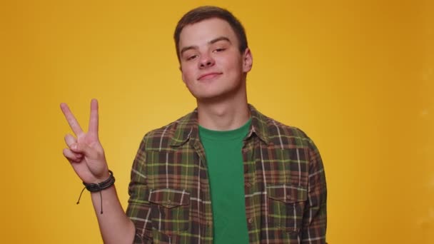 Teen man in casual green shirt showing victory sign, hoping for success and win, doing peace gesture, smiling with kind optimistic expression. Young adult guy boy isolated on yellow studio background - Footage, Video